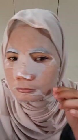 Here is a description of the video:
A person wearing a light pink hijab is applying a sheet mask to their face. The mask is white with blue accents around the eye area. The person uses their fingers to smooth out the mask around their eye, nose, and mouth, ensuring it adheres to their skin. The background is a plain white wall with what appears to be a white cabinet door visible. A key and a silver clip hang on the wall behind the person.