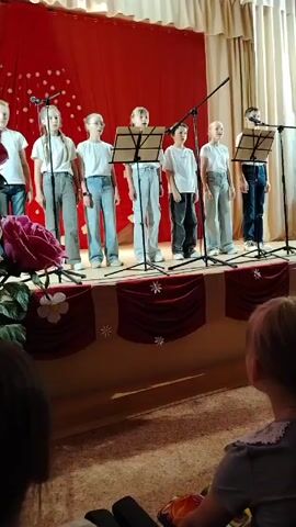 Seven children, wearing white t-shirts and jeans, are standing on a stage with a red curtain backdrop adorned with white snowflake designs. Each child stands behind a microphone and music stand. The stage is decorated with red fabric featuring more snowflake designs. In the foreground, a person is sitting and watching the performance. A large floral arrangement is visible on the left side of the stage.