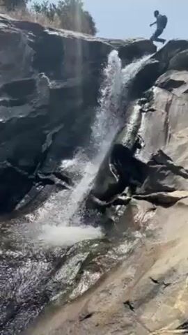 The video shows a waterfall cascading down a rocky cliff face. Three people are visible at the top of the waterfall, appearing to be preparing to jump into the pool below. The environment is sunny and bright, suggesting a clear day. The rocks surrounding the waterfall are a mix of dark and light colors, indicating various mineral compositions or levels of moisture. The water is clear and splashes as it hits the pool.