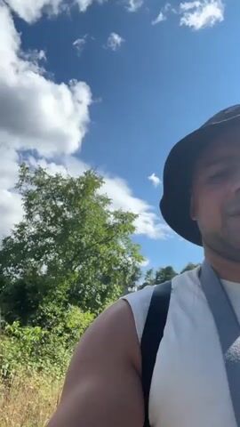 The video begins with a shot of a paved path winding through a green, wooded area on a sunny day. The camera then pans to reveal a man wearing a white t-shirt, a bucket hat, and a backpack. He is walking on the path and looking at the camera. The background remains a mix of trees, vegetation, and a bright blue sky with white clouds.