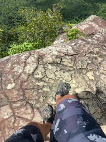 The video begins with a close-up of a person's legs and feet standing on a textured, rocky surface. The person is wearing dark camouflage shorts and dark perforated sandals. The camera then pans up and out, revealing a vast, lush green landscape stretching out towards a blue ocean under a partly cloudy sky. The viewpoint is from a high elevation, likely a mountaintop, overlooking a jungle, a winding river, and a coastal town. Several islands are visible in the distance. The sun casts shadows across the landscape, indicating it is daytime. The overall scene depicts a scenic and natural environment.