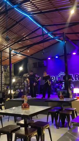 A live band performs on a stage in a dimly lit, indoor venue. The stage is illuminated by purple and blue lights, with a neon blue light strip running along the ceiling. The band consists of at least four members: two singers at the front, a guitarist, and a drummer. The singers are dressed in black, with one female singer wearing a black top and pants with a white sash, and a male singer in a black t-shirt and pants. Behind the band, a large sign with the text "PILLAR BAR" is visible. In the foreground, there are several tables with marble-like tops and dark stools, suggesting a bar or restaurant setting. The overall atmosphere appears to be lively and entertaining, with the band actively engaged in their performance. The time of day is likely evening or night due to the indoor lighting and performance.