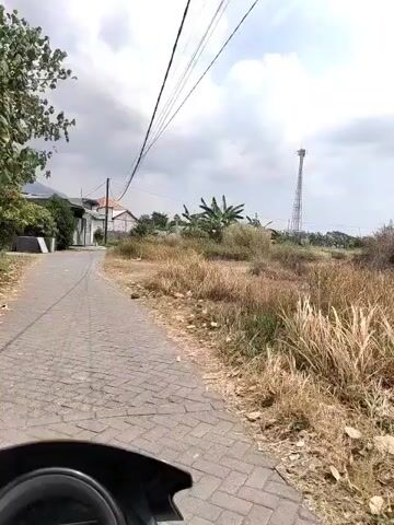 The video captures a point-of-view shot from a moving vehicle, likely a motorcycle, on a paved road with cobblestone-like patterns. The road curves to the left, bordered on the right by a dry, overgrown area with tall, brown grass and scattered debris. In the distance, a tall cell tower is visible. To the left of the road, there are houses with white walls and red-tiled roofs, and some trees and vegetation. The sky is overcast with visible power lines stretching across the frame. The overall scene depicts a rural or semi-rural environment on a cloudy day.