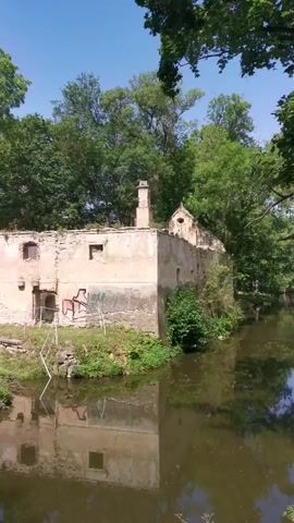 The video shows an abandoned building next to a body of water. The building appears to be made of stone and has a crumbling facade with visible graffiti. Trees surround the building and water, with their reflections visible on the water's surface. The sky is clear and blue.