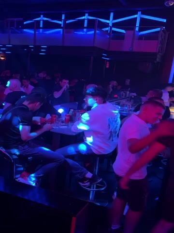 The video shows a nightclub with people gathered around tables and standing. The atmosphere is dark with blue and pink lighting, and some spotlights are visible. Several individuals are interacting with each other and with drinks on the tables. The camera pans across the room, offering glimpses of the crowd and the club's interior design, which includes illuminated structures on the walls.