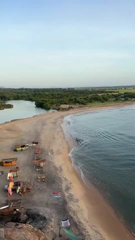 This aerial video captures a serene beach scene in Shastrawela, Sri Lanka, during what appears to be the early morning or late afternoon, given the soft, diffused light and clear sky. The primary subjects are the pristine sandy beach, the gentle turquoise ocean with small waves lapping at the shore, and a lush green treeline bordering the sand, suggesting a tropical environment.

On the beach, several rustic wooden stalls or stands are set up, displaying various surfboards and what might be local crafts or merchandise. A few people are visible: one individual walks along the shoreline, and two others are near the water's edge, possibly engaging in water activities. A small, light-colored vehicle, possibly a tuk-tuk or a small van, is parked among the stalls, adding a touch of local life. Further inland, a calm body of water, likely a lagoon or river, is visible, contrasting with the dynamic ocean.

The overall mood is peaceful and tranquil, emphasizing the natural beauty of the location. The weather seems pleasant and calm, suitable for beach activities. No text is visible in the video.