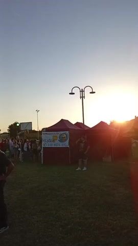 The video depicts an outdoor gathering at sunset. In the foreground, on the left, a man in a dark shirt and pants stands with his hands on his hips, facing left. To the right of the man, a tent with a purple canopy and a sign featuring the word "Mister POP" is visible. Behind the tent, a line of people is forming. Further in the background, an inflatable blue archway with the word "AVIS" on it spans a grassy area, with more people gathered around it. The sun is setting on the right side of the frame, casting a warm glow over the scene. Several lampposts are scattered throughout the area. The ground is covered in grass. The sky is clear and blue. People are dressed in casual summer attire. Some individuals appear to be talking to each other, while others are walking or standing in line. The overall atmosphere suggests a community event or festival.