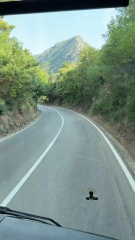 The video shows a POV perspective from inside a vehicle, likely a bus or van, as it navigates a winding road through a mountainous, forested area. The road is paved and has a solid white line marking the edge and a dashed white line in the center, indicating a two-lane road. The surrounding landscape is lush green with dense trees lining both sides of the road, and a mountain range is visible in the background under a clear, bright sky, suggesting a sunny day.

In the distance, on the left side of the road, a red vehicle is visible. Further ahead, there are road signs, including a triangular warning sign with a red border and a symbol, possibly indicating a sharp curve or a hazard. There are also some smaller structures or buildings nestled into the hillside on the right side of the road. A convex mirror is mounted on a pole on the right, presumably to aid visibility around a blind curve.

The overall impression is of a scenic drive through a natural landscape, possibly in a rural or mountainous region. The sunlight and clear sky suggest pleasant weather. The presence of the road, signs, and mirror indicates that this is a traveled route. The camera is mounted inside the vehicle, and a small black clip-like object is visible on the dashboard in the lower center of the frame. No people are directly visible in the video, but the activity is clearly driving. The location is identified as Mišići, Montenegro.