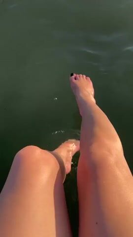 The video shows a pair of legs dangling in murky green water, with the feet occasionally splashing. The sun appears to be shining, casting a warm glow on the skin. The scene suggests a leisurely activity, perhaps swimming or sitting by a body of water on a warm day. The location is identified as Verbizh, Ukraine. There is no visible text in the media file.