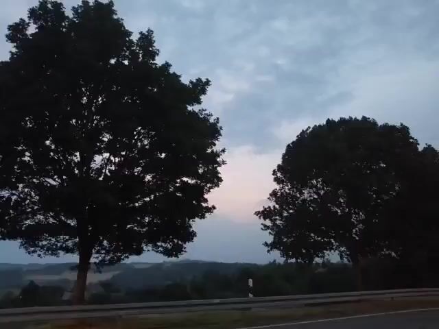 The video shows a view from a vehicle moving along a road. Two large trees are visible in the foreground, silhouetted against a cloudy sky at dusk. The sky is a mix of blue and gray, with hints of pink and orange near the horizon. Beyond the trees, a rolling landscape with hills and scattered vegetation can be seen. The road appears to be paved, and a metal guardrail runs along its edge. The vehicle's headlights are on, casting a faint light on the road surface. The overall mood is peaceful and serene.