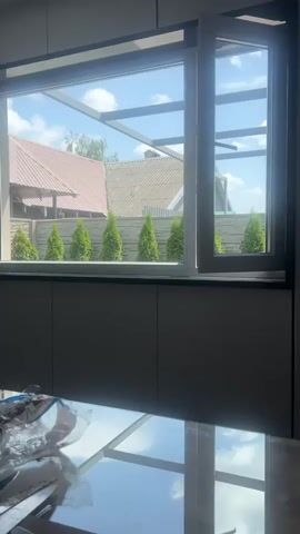 The video shows a partially opened window with a view of a backyard. Outside the window, there are several evenly spaced evergreen trees. Behind the trees, a fence and the roof of a building are visible. The sky is blue with some clouds. The interior of the room shows a countertop and part of a table with a reflection of the sky and clouds. The window frame is black and white.