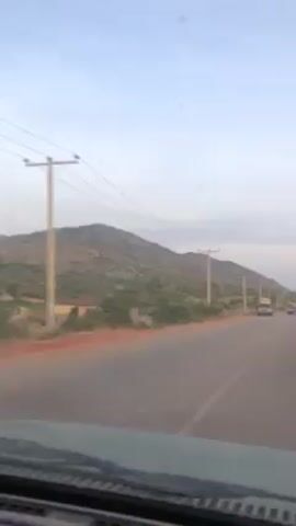 The video shows a drive on a paved road through a rural landscape. In the initial shot, the road stretches ahead toward a large, rocky mountain or hill. Power lines run alongside the road. There are a few buildings visible on the left, and a few cars are driving in the opposite direction. The subsequent shots provide a clearer view of the landscape. The area is dry, with sparse vegetation and rocky hills in the background. The sky is overcast. The road is mostly empty except for the car the footage is taken from.