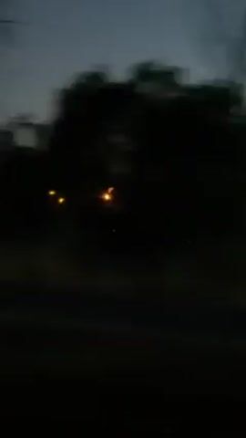 The video shows a dark landscape at dusk or night. There are lights from a factory or industrial complex visible in the distance. The camera is moving, likely from a vehicle, as trees and other objects in the foreground pass by quickly. The sky is a dark gradient, suggesting the end of daylight. There is no indication of people or any specific activity beyond the lights of the industrial facility.