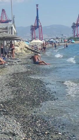 A person sits on a pebbled beach with their legs in the shallow water. The waves gently lap at the shore. Behind them, other beachgoers are scattered along the sand and in the water. In the background, large blue and red cranes dominate the skyline, suggesting a port or industrial area nearby. The sky is clear and the sun is shining.
