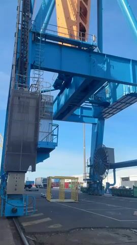 The video shows a large blue and yellow crane next to a cruise ship. The crane has a lift and a ladder attached to it. The cruise ship is white with many windows, and there is scaffolding around some of the windows. There is a gangway attached to the cruise ship. There are workers on the gangway and on the scaffolding of the ship. The sky is blue.