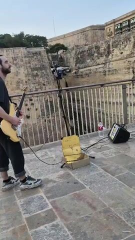 A man with dark hair and a beard is playing a light-colored electric guitar. He is wearing a dark t-shirt and dark pants rolled up at the ankles, with patterned sneakers. He stands on a stone-paved walkway with a metal railing to his right. Behind him is a tall, ancient stone wall. In front of him, on the ground, is a makeshift donation box made from a cardboard box. A microphone stand with a microphone is also set up, along with a speaker and a bottle of water. The background shows more of the stone wall, trees on top of it, and a cityscape with buildings and a cloudy sky. The video pans slightly to the right, revealing a person's legs and feet in the lower right corner of the frame.