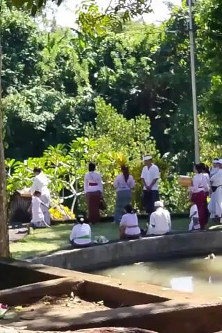 A group of people, some dressed in white tops and sarongs with white headbands, are gathered near a body of water. Several individuals are seated facing the water, while others stand behind them. One person is holding a basket. The environment appears to be outdoors with lush green trees in the background. The water has lily pads and some movement visible on its surface. The lighting suggests a sunny day.