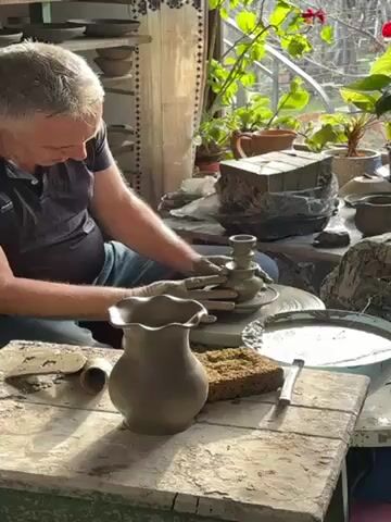 The video features a gray-haired man seated at a pottery wheel, shaping a clay object. He is using his hands to mold the clay, which appears to be a candlestick holder. The wheel is spinning as he works. In front of him on a wooden table is a finished vase with a ruffled edge, a sponge, and a tool. To his left, there is a shelf lined with pottery pieces. Behind him, a window provides natural light, illuminating various potted plants and objects in the background. The setting suggests a pottery studio or workshop.