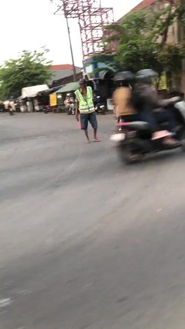 The media file depicts a street scene in Juwet Kenongo, Indonesia. The primary subject is a person wearing a reflective vest and holding a red baton, likely directing traffic. Another individual on a scooter, blurred by motion, is also visible. The setting appears to be a street with buildings and trees lining the background. The time of day seems to be late afternoon or early evening, as indicated by the lighting and the sky. The weather appears to be clear. There is a sign with yellow text visible in the background, but the text itself is illegible due to the distance and resolution of the image. The overall mood of the image is that of an active street, possibly experiencing some traffic management.