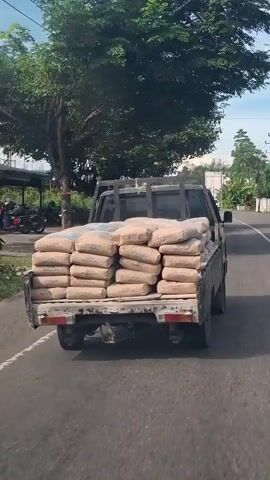 A pickup truck is driving down a paved road. The truck bed is filled with stacks of bagged cement. Trees and vegetation are visible on the left side of the road, and on the right, there is a road with other vehicles and utility poles. The sky is partly cloudy.