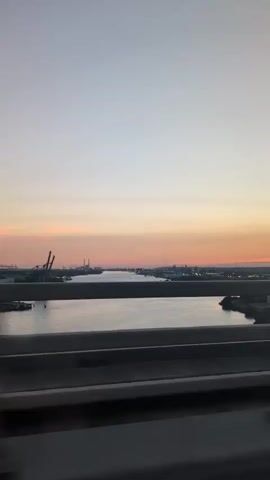 The video shows a view from a moving vehicle, likely a car, looking out over a wide body of water. The water reflects the sky, which is a pale gradient of soft blues, pinks, and oranges, suggesting either sunrise or sunset. In the distance, across the water, is a cityscape with industrial elements, including several large cranes and buildings. The water itself appears calm, with a few small boats visible. A concrete barrier or railing is in the foreground, blurring the view slightly and indicating the perspective of someone inside a vehicle. The overall atmosphere is tranquil and scenic.