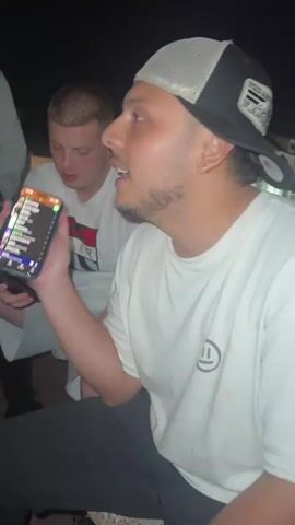 Two young adult males are outdoors in dimly lit conditions, possibly at night. The male in the foreground is wearing a white t-shirt with a smiley face logo and a black baseball cap with a mesh back. He is holding up a smartphone, displaying a colorful list of names and numbers, and appears to be speaking. He is looking to his right, out of frame. The second male is seated behind and to the left of the first, wearing a white t-shirt with red lettering. He is looking down at a smartphone in his hands. The background is dark and out of focus, with faint hints of outdoor structures or lights.