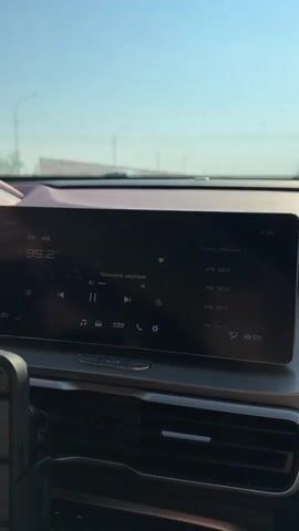 The video shows the interior of a car, focusing on the dashboard with a large digital display screen. The screen shows radio station information, with the current station at 95.2 FM. There are playback controls on the screen, including pause and play buttons. The time is displayed as 00:00. The dashboard also shows the temperature outside, which is 18.5 degrees Celsius. The view outside the car through the windshield shows a clear sky and a road with a barrier on the side. A smartphone is mounted on the left side of the dashboard.