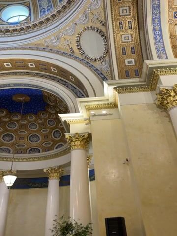 This media file captures the interior of a grand, ornate dome, likely a place of worship or a significant public building. The dome is richly decorated with intricate patterns in gold, blue, and white. Sunlight streams in through several circular windows embedded in the dome's upper section, suggesting it is daytime.

The dome's interior is a complex architectural marvel. Bands of geometric patterns, possibly of a diamond motif, circle the apex. Smaller domes or alcoves with celestial-like blue ceilings adorned with circular patterns are visible. A large, elaborate golden chandelier hangs from the center, further accentuating the opulence of the space. White, fluted columns topped with golden Corinthian capitals support the surrounding architecture, adding to the sense of grandeur.

The overall impression is one of solemnity and majesty. The lighting, primarily from the windows and the chandelier, creates a warm and inviting atmosphere within the vast space. While no people are visible, the presence of a large speaker at the bottom right suggests that an event may be taking place or has recently concluded, potentially a concert, ceremony, or public gathering. The architectural style hints at a historical building, possibly with religious or governmental significance, given the scale and decorative elements. The location context provided, посёлок дачного хозяйства «Архангельское», Russia, places this impressive structure within a specific geographical area, known for its historical estates and cultural significance.