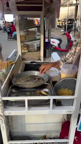 The video shows a street food vendor in Rhieng Blang, Indonesia, preparing fried snacks. The vendor, a woman wearing a hijab and a plaid shirt, is actively engaged in cooking. She is using a cart equipped with a deep fryer, a metal grate for draining oil, and containers for ingredients. Skewered food items are being fried in a large pan. To her right, a strainer holds a pile of shredded ingredients, likely for coating or serving with the fried items. A clear pitcher filled with a yellow liquid, possibly batter, is also visible.

The background depicts a street scene with other food stalls and activity. Scooters are parked nearby, and people are walking or riding on them. The lighting suggests it is daytime, possibly late afternoon or early evening, given the warm, diffused sunlight. The overall atmosphere appears to be a typical bustling street market.

Visible text on the red canopy above the cart reads "NUGGET GORENG" and "TAHU GORENG," indicating that fried nuggets and tofu are among the items being sold. The scene is lively and authentic, capturing a common aspect of Indonesian street food culture. The vendor's focused action highlights the preparation process of the food being offered to customers.