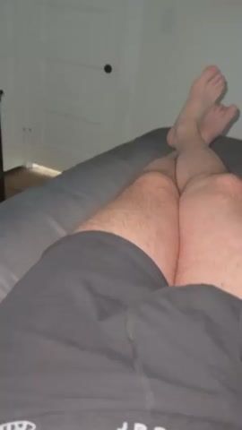 This is a close-up, handheld video recording from a first-person perspective, showing a person's legs and feet as they lie down. The person is wearing dark shorts, and their legs are hairy. Their feet are crossed at the ankles and are positioned at the top of the frame, with the toes pointing upwards.

The background suggests an indoor setting. To the left, a white door with a dark doorknob is visible, possibly indicating a bedroom or a similar private room. A wall-mounted television is visible on the right side of the frame, turned off. Above the television, a black ceiling fan with three blades is in motion, suggesting the presence of air circulation. The lighting is dim, creating a twilight or evening atmosphere.

There is no visible text in the video. The overall impression is one of relaxation or repose in a private living space, likely at home in Des Moines, United States of America, during the evening.