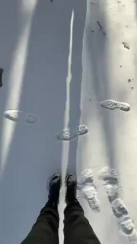The video shows a person standing in the snow looking up at the inside of a cooling tower on a sunny day. The camera is positioned looking down at the person's boots, and as the camera pans up, the person's shadow is cast on the snow in front of them. The shadow is long and thin, and there are footprints in the snow. The person is standing inside a large, circular cooling tower. The tower is made of concrete and has a large hole at the top, through which the blue sky can be seen. There are metal beams and pipes running across the inside of the tower. The person is wearing a dark jacket and pants.