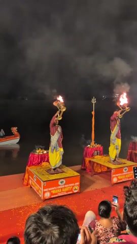 The video captures an outdoor evening ceremony. Two men in traditional Indian attire, maroon shirts and yellow sarongs, stand on elevated platforms decorated with red fabric and flower petals. Each man holds a brass lamp with a flame, performing a ritualistic dance or offering. Smoke billows from the lamps and around them. In the background, a dark body of water is visible, with distant lights suggesting a city or town. Several people are seated in the foreground, facing the performers and capturing the event with their phones. The overall atmosphere is one of reverence and cultural performance.
