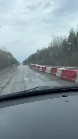 The video shows a view from inside a car driving on a heavily damaged road through a forest. The road is wet and has numerous potholes and patches of missing asphalt. A long line of white and red plastic barriers runs along the right side of the road. A few orange traffic cones are also visible. The sky is overcast, and there are signs of light rain on the windshield. Bare trees line both sides of the road, indicating it is likely late fall or early spring. Another vehicle is visible in the distance. The focus is on the poor road conditions and the traffic control measures in place.