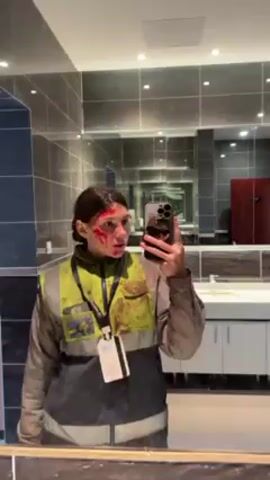 A woman stands in a bathroom, taking a selfie in the mirror. She has what appears to be fake blood on her face. She is wearing a reflective work vest and a lanyard. The bathroom has gray tiled walls, recessed lighting, and a white sink. A red door is visible behind her in the mirror's reflection. The woman is holding a cell phone in her right hand. She appears to be saying something.