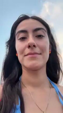The video shows a woman standing on a beach. She is facing the camera and smiling slightly. She has dark hair and brown eyes, and is wearing a red floral dress with a blue bathing suit top underneath, as well as a gold necklace and yellow stud earrings. The beach and ocean are visible behind her, and the sky is a light blue with some clouds. Other people can be heard talking in the background.  A hand comes into frame on the right at about the 3 second mark.