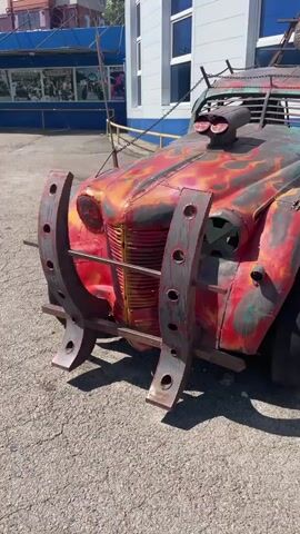 The video shows a close-up, panning shot of a heavily modified, post-apocalyptic-style truck parked on an asphalt surface. The truck has a distressed, rusty appearance with flames painted on the hood. It features a large, exposed engine intake, metal bars covering the grille and windows, and what appear to be spikes on the roof. Large, U-shaped metal bumpers are attached to the front. The background shows a building with blue awnings and a fence. The lighting suggests it is daytime with bright sunlight casting shadows. No people are visible in the frame.