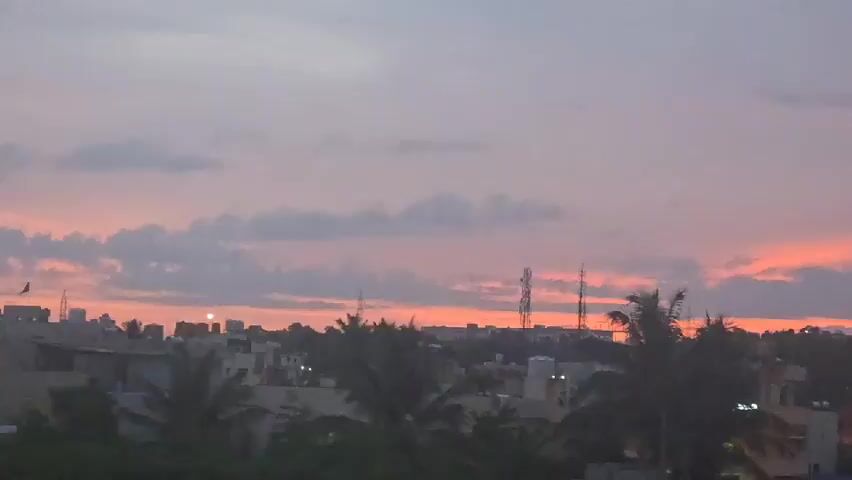 A time-lapse video shows a dusky sky over a city skyline. The sky transitions from a pale purple to a deeper purple and orange. Silhouetted buildings and trees are visible against the sky. Several tall, thin towers, possibly cell towers, are visible in the distance. Streetlights and lights from some buildings illuminate the scene. The camera pans slightly to the right, capturing more of the city and sky. The video ends with the sky becoming darker and the lights of the city becoming more prominent.