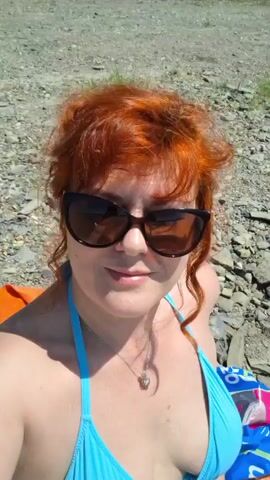 A woman with red hair and sunglasses is lying on the beach, wearing a blue bikini. She is taking a selfie and making faces. The beach is made of stones and there are some towels in the background.