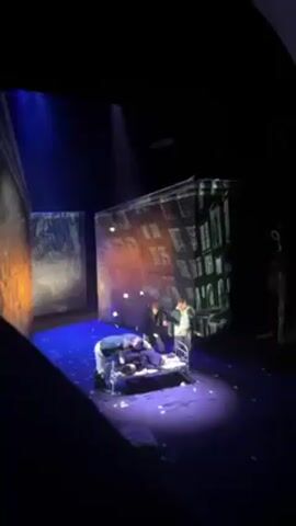 The video shows a scene from a theater production. Five men stand around a metal-framed bed. One man is lying on the bed, while the other four men are leaning over him, looking down. The stage is lit with a blue light and a spotlight focused on the bed. The set includes backdrops with illustrations of buildings and a tree. The floor is also littered with small white pieces of paper.