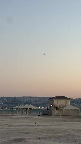 A vertical, medium shot shows a distant airplane flying in a clear, pale sky over a sandy beach. A few people walk on the beach near white beach huts and gazebos. In the far background, a city skyline with buildings and lights is visible. The water of the sea is between the beach and the city. The sky is clear, suggesting a sunset or sunrise, with a light gradient from pale blue to light pink.