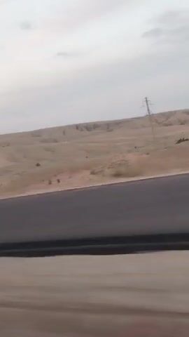 This media file captures a landscape view from a moving vehicle, likely a car, on a road. The scene is dominated by vast, arid, and hilly terrain with a muted color palette. The ground appears dry and dusty, with patches of sparse vegetation and some signs of human activity, such as what looks like agricultural terracing on the hillsides. A prominent feature in the distance is a tall, lattice-style electricity pylon, indicating the presence of power infrastructure. There are also some man-made structures visible, including a road with clear lane markings and a yellow and black diagonal warning sign on the side of the road, suggesting a curve or hazard.

The time of day appears to be either early morning or late evening, as the sky is overcast with soft, diffused light and some wispy clouds, with hints of pink suggesting sunrise or sunset. The weather seems calm, with no strong winds evident. The overall mood is one of a journey through a remote, sparsely populated, and somewhat desolate landscape.

The location is identified as Mukry, Kazakhstan. There is no discernible text in the media file, aside from what might be on the warning sign, which is too blurred to read. No people are clearly visible in the frame, only implied by the presence of the road and infrastructure. The media file is a series of short video clips, each showing a slightly different perspective of the same scenery as the vehicle progresses.