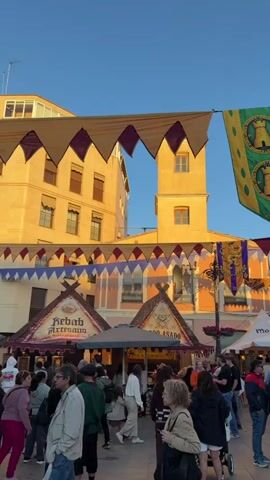The video shows a town square with a market, possibly a medieval festival. Numerous people, including adults and children, are walking around, browsing the stalls, and interacting with each other. Above the crowd, strings of triangular flags, alternating between yellow, red, and purple, are strung between the buildings. There are stalls selling food, including “kebab artesano” and “asado.” A green and yellow banner featuring a castle tower hangs in the foreground. The buildings around the square are a mix of colors, with some being yellow, pink, and red brick. The sky is a clear blue.