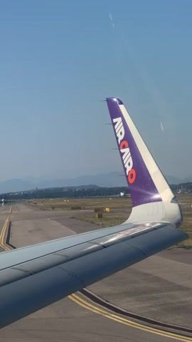 The video shows the tail fin of an Air Cairo airplane, which is purple and white with the airline's logo. The airplane is on a runway, with other aircraft and airport infrastructure visible in the background. The sky is clear and blue, suggesting it is daytime. The camera is looking out of an airplane window, showing a perspective of the wing and the runway. The airplane appears to be taxiing or preparing for takeoff.