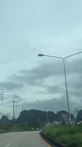 This media file depicts a cloudy day in Ao Luek, Thailand, viewed from a moving vehicle. The scene is dominated by a road winding through a lush, green landscape. Towering, jagged limestone karsts covered in vegetation rise majestically in the background, characteristic of the region's geography. The sky is overcast with heavy, gray clouds, suggesting a potential for rain or a recent downpour.

Along the roadside, electrical poles with visible wires stretch across the frame, hinting at the presence of infrastructure. Several streetlights, designed for roadways, are also prominent, some illuminating the scene despite the dim light. In the middle ground, a line of buildings and homes can be seen nestled amongst the greenery, indicating a settled area or small town. The road appears to be a moderately busy thoroughfare, with a few vehicles visible in the distance.

The overall impression is one of a somewhat subdued, yet naturally beautiful environment. The cloudy weather creates a slightly dramatic or moody atmosphere, contrasting with the vibrant green of the foliage and the imposing, ancient feel of the karsts. The perspective from within a vehicle implies travel, a journey through this scenic and geologically unique part of Thailand. No people are explicitly visible, but their presence is implied by the infrastructure and the road. The time of day is difficult to pinpoint precisely due to the overcast conditions, but it appears to be daytime. There is no discernible text within the frame.