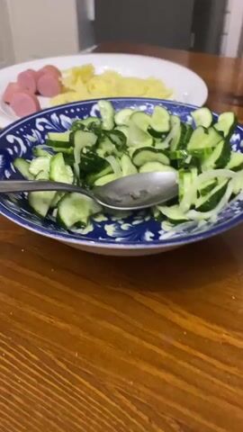A close-up, slightly shaky shot shows a plate of sliced cucumbers and onions in a blue and white patterned bowl. A metal spoon rests in the bowl. In the background, on a white plate, are sliced sausages and pasta. The food is on a wooden table.