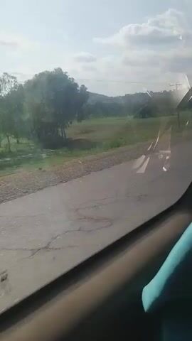 The video shows a view from inside a vehicle looking out the side window. The vehicle is moving along a paved road. On the right side of the road, there is a grassy area with some trees and rolling hills in the distance. The sky is partly cloudy. There is a utility pole visible on the right side of the road. The interior of the vehicle is visible in the foreground, including a part of the car door and window frame.