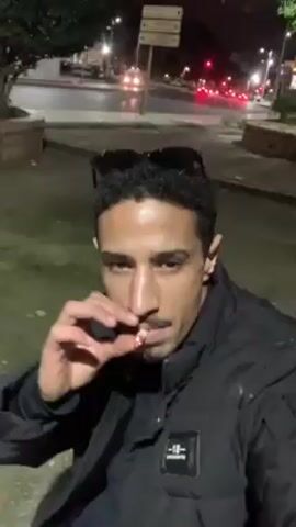 The video shows a man smoking what appears to be a cigarette or similar roll-up outside at night. He is wearing a black puffer jacket, and sunglasses are perched on his head. The background reveals a street scene with lights, traffic, trees, and some low walls. The man is looking at the camera and exhaling smoke. The area seems to be a small park or green space near a roadway.