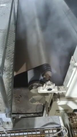 The video shows a piece of industrial machinery, including a wide metal belt and a series of rollers and other components. Vapor or steam is visible around the machinery, suggesting the presence of heat or a humid environment. The machinery is located on a platform with metal grating. The camera is positioned at an elevated angle, capturing the moving belt and various parts of the machine. The lighting is dim, typical of an industrial setting. No people are visible in the video.