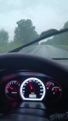 This is a first-person view from inside a car on a rainy day. The windshield wipers are active, clearing raindrops from the view of the road ahead. The dashboard is visible, showing the speedometer indicating a speed of approximately 100 km/h. The road appears to be a two-lane highway surrounded by trees. The sky is overcast and the visibility is reduced due to the weather.