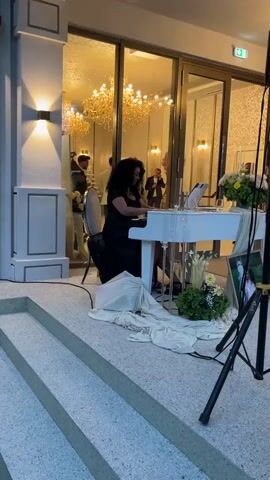 A woman in a black dress plays a white piano in front of a building with large glass doors. Inside the building, chandeliers are visible. A man in a white jacket stands and talks on a phone, and another person in a dark dress stands near him. Outside, a large speaker on a stand is to the right of the piano, and a white cloth is draped over a box or stand in front of the piano. Two steps lead up to the area where the piano is located.